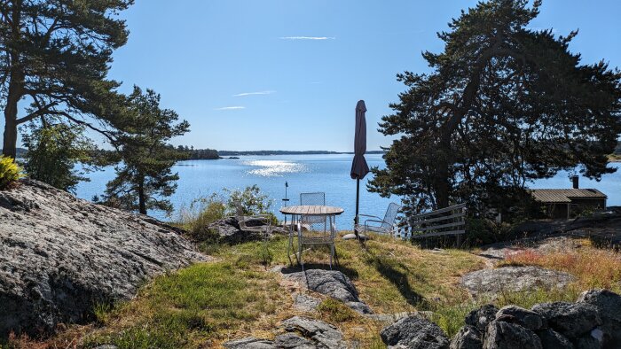 Trädgårdsbord och stolar med utsikt över solbelyst sjö, omgivna av träd och klippor.