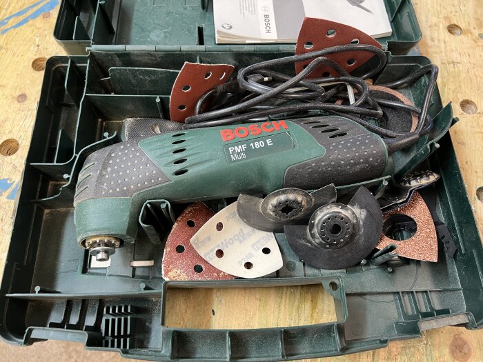 Bild på ett multitool från Bosch med modellnamnet PMF 180 E Multi och tillhörande verktyg och slippapper i en öppen förvaringslåda.