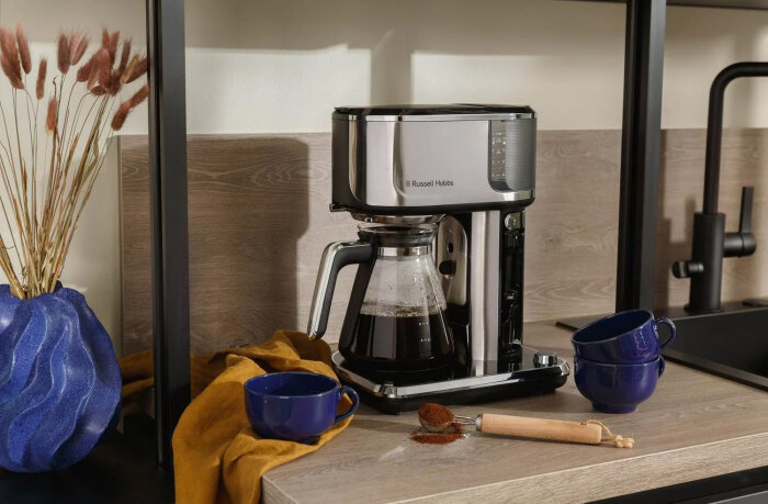 Russell Hobbs kaffebryggare på en köksbänk med blå vas, två blå koppar, gul handduk och kaffepulver med doseringsskopa intill.