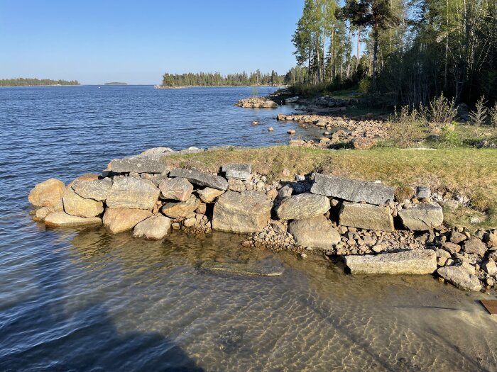 Gammal stenpir vid vattenkant, delvis under vatten, med skog i bakgrunden, norr om Gävle. Stora stenar och gräsmark ovanför piren.