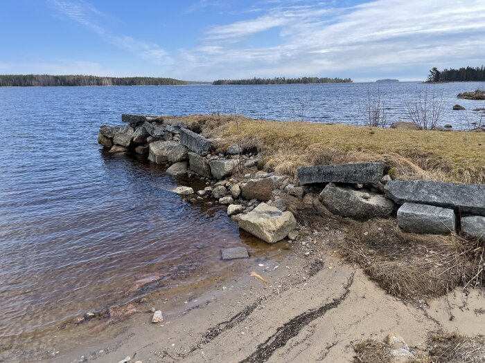 Gammal stenpir vid en skyddad vik med utsikt över vatten och skog i bakgrunden, planerad plats för brygga ovanpå piren.