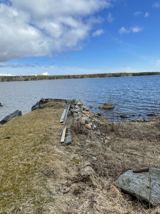 En gammal stenpir med några plankor ovanpå som sträcker sig ut i en sjö, omgiven av gräs och stenar, med skog och blå himmel i bakgrunden.