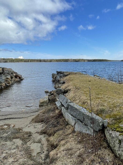 Stenpir vid vattenkant omgiven av gräs och grus, med utsikt över ett lugnt vatten och en skog i bakgrunden under en delvis molnig himmel.