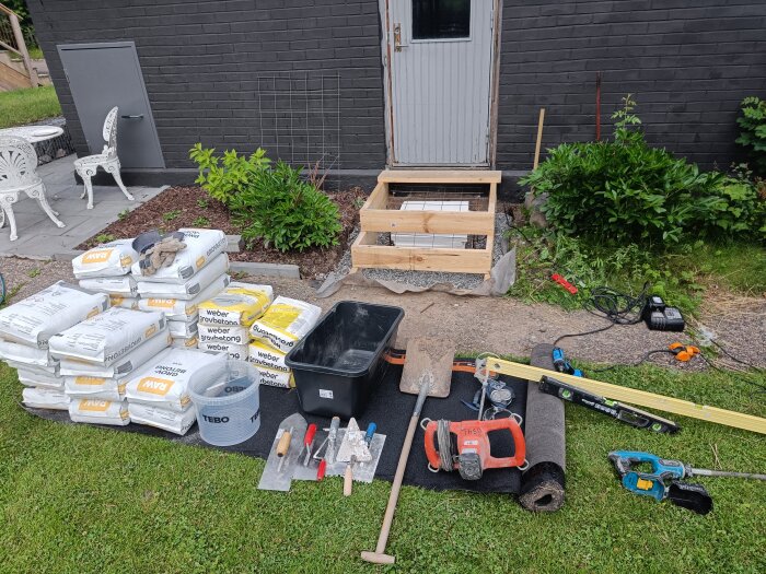 Arbetsstation med byggmaterial, verktyg och betongblandning framför en trappa i träkonstruktion, bredvid en dörr och växter.