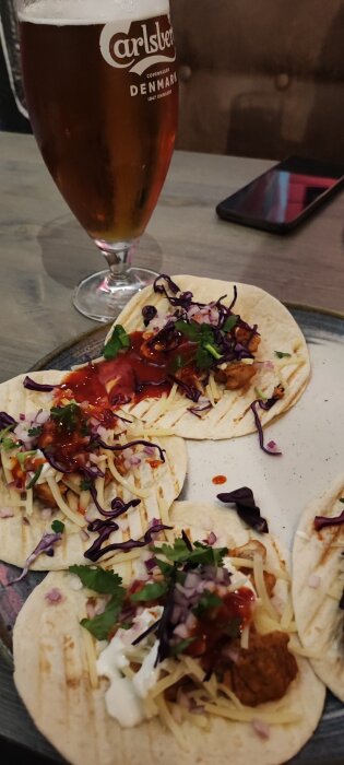 Tacos med kött, lök, koriander, salsa, och vit sås samt ett glas öl på ett bord. Bakom glaset syns en mobiltelefon.