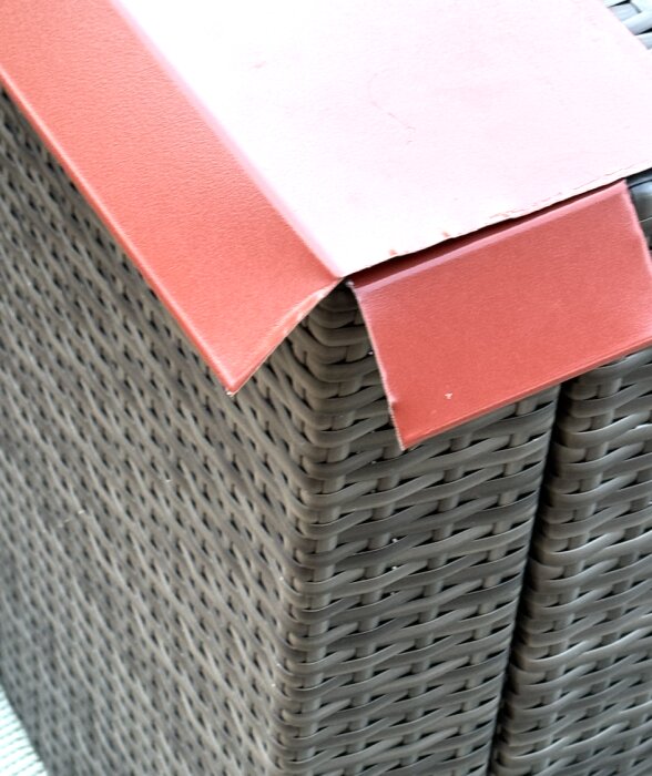Närbild av en hörnbit där en rödaktig plast- eller metallrem fogats mot hörnet på en stupränna av vävd grå plast, med en öppen triangelhålighet.