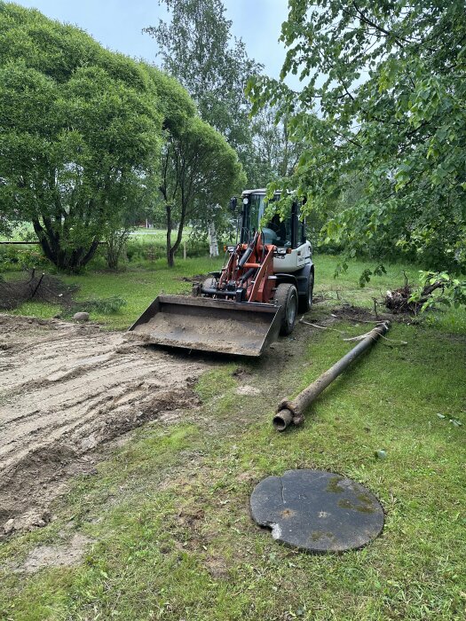 Traktor på gräsmatta framför träd, som används för att flytta gamla vattenrör efter avloppsrörsbyte. Ett vattenrör och ett brunnslock ligger på marken.