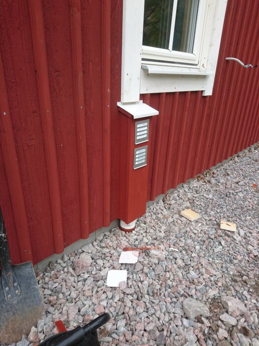 Röd husvägg med vitmålat fönster och en tilluftskanal monterad under. Tilluftskanalen är inkopplad till ett rör som leder ner i stenkross vid husets grund.