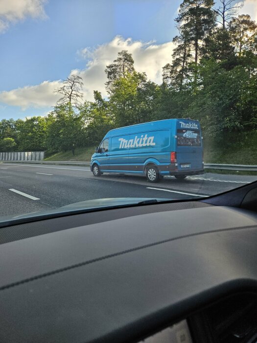 En blå Makita-skåpbil kör på en motorväg, omgiven av grönskande träd och klar himmel i bakgrunden.