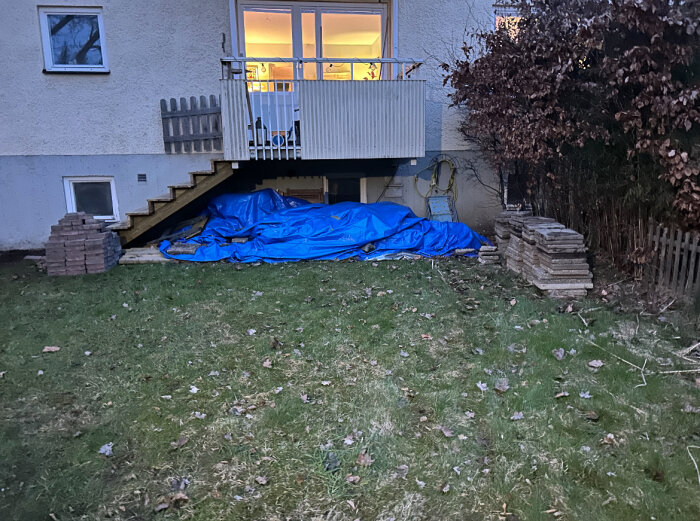 Baksidan av en bostadsrätt med en presenning under balkongen och staplade stenar på sidorna, tagen i skymningen med gräsmatta i förgrunden.