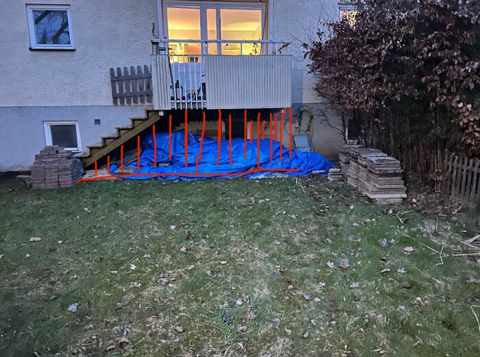 Blå presenning täcker marken under en balkong på en lägenhet i Stockholm; tegelstenar staplade på båda sidor, marken är grästäckt med löv.