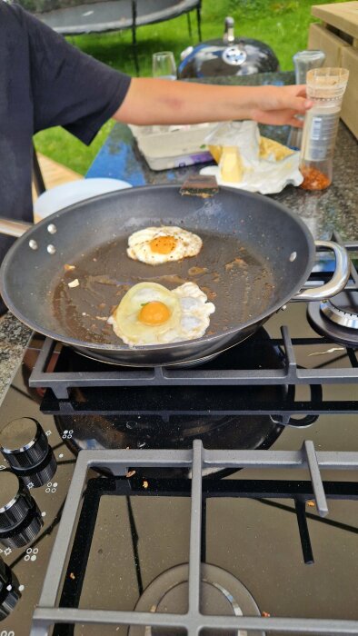 Person steker ägg i en stekpanna på en gashäll i ett utomhuskök, omgiven av olika matvaror och redskap.