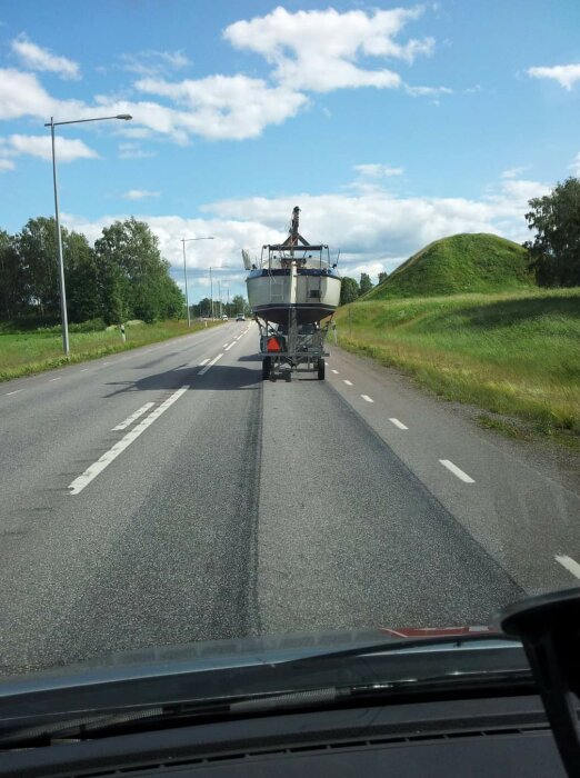 En bagagevagn som används på flygplatser transporterar en båt på en landsväg med gräs och kullar i bakgrunden.