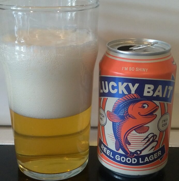 En öl i ett glas bredvid en läskburk med etiketten "Lucky Bait Reel Good Lager".