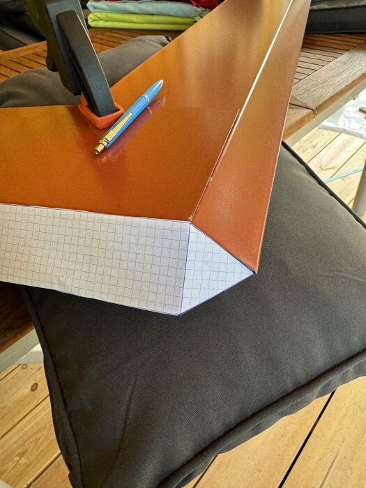 En pappersskiss av ett hörn på en takdel i metall med en blå penna intill, placerad på kuddar och en bordsyta.
