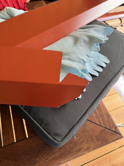 Handskklädda händer håller två hörn av ett rött plåtstycke med triangulära utskärningar, på en kudde som ligger på ett träbord.