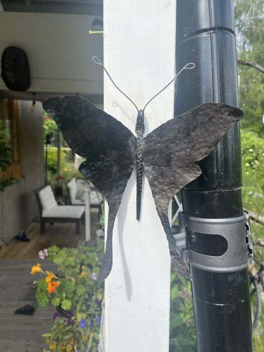 Självgjord fjäril i smide fastsatt på en vit stolpe vid en uteplats med gröna växter och blommor i bakgrunden.