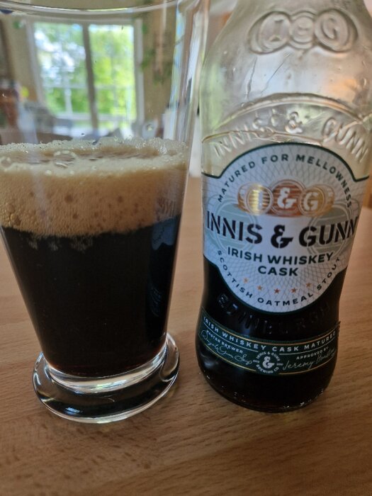 Flaska och glas med mörk öl, märket Innis & Gunn Irish Whiskey Cask, på ett träbord. Bakgrunden visar ett suddigt fönster med träd utanför.