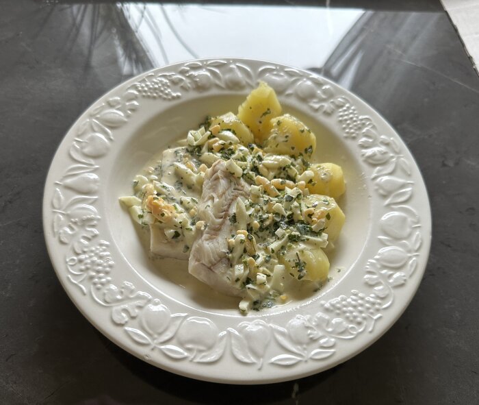 En tallrik med kokt fisk, potatis och en krämig sås med hackad persilja och äggbitar, serverad på ett grått bord.