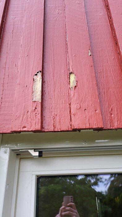 Röd träpanel på husgavel med flagnande färg och synliga skador på träet ovanför en fönsterram.