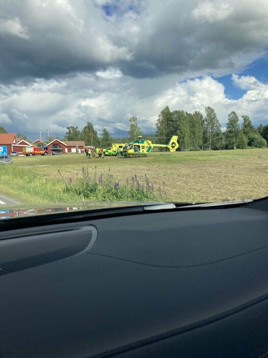 En gul räddningshelikopter och ambulans står på en åker med rödvita byggnader och en brandbil i bakgrunden, omgivna av träd och molnig himmel.