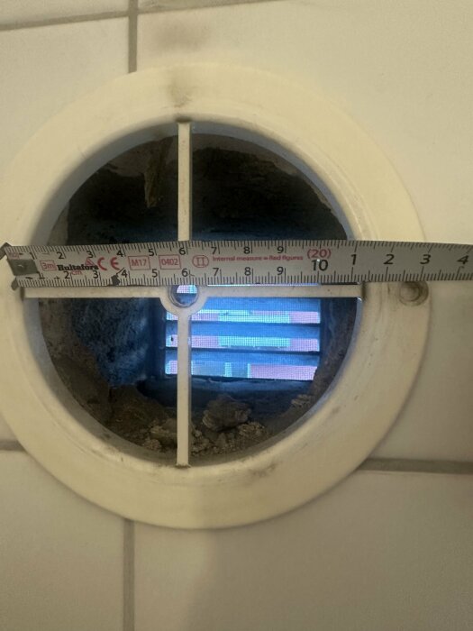 Närbild på en rund ventil i badrumsvägg med måttband som visar diametern som ca 115mm.