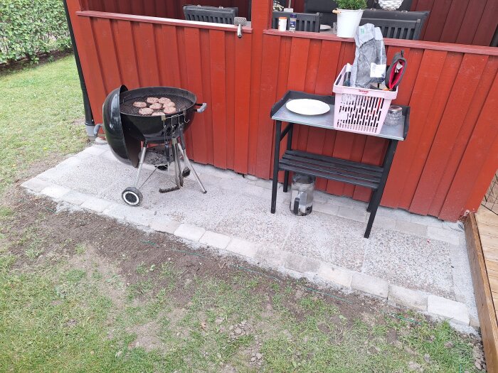 Svart klotgrill med hamburgare på en uteplats av stenplattor framför en röd trävägg. Brevid grillen finns ett svart metallbord med en korg och tallrikar.