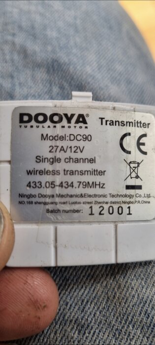 Närbild på en fjärrkontrolls baksida med etikett som visar: Dooya Tubular Motor, Model: DC90, 27A/12V, Single channel wireless transmitter, frekvenser, och batchnummer 12001.