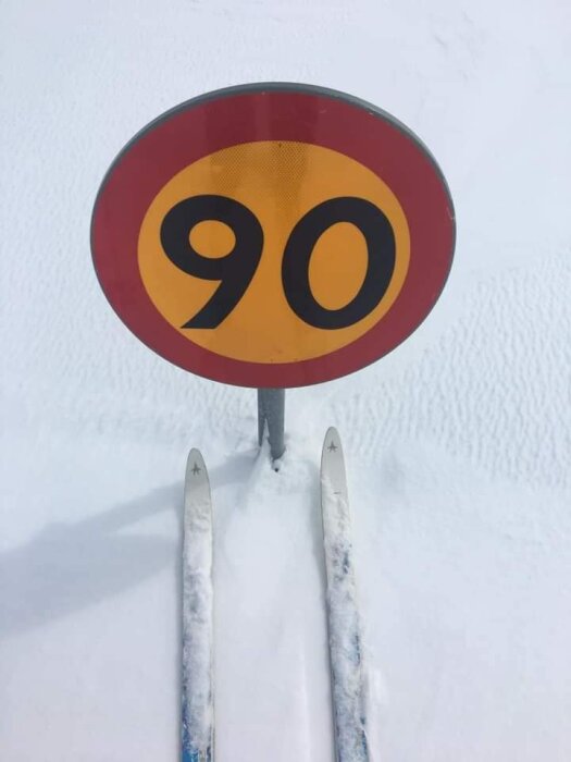 Två skidor framför en fartbegränsningsskylt med texten "90" i snötäckt landskap.