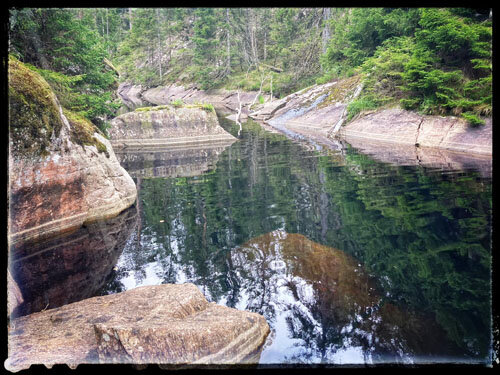 En skogstjärn omgiven av klippor och tät skog. Vattnet är klart och reflekterar de omgivande träden och klipporna.