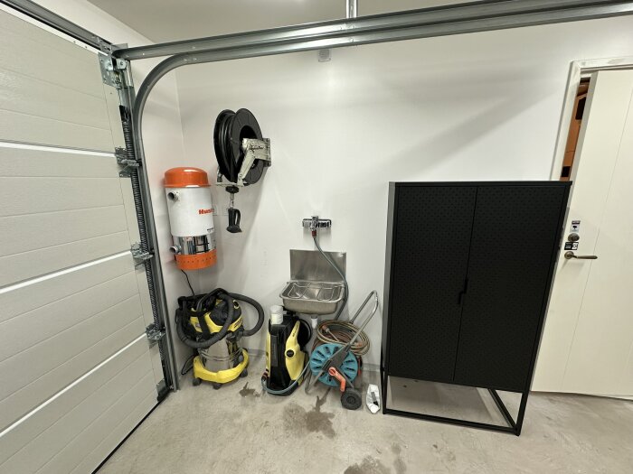 Garage med centraldammsugare, högtryckstvätt med slangvinda på väggen, förvaringsskåp, ho samt en dammsugare på golvet nära en garageport.