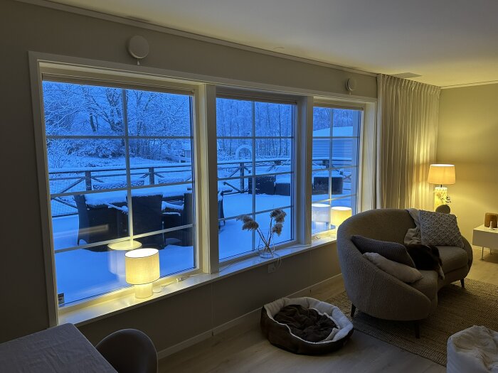 Vardagsrum med tre stora fönster mot en snötäckt altan, soffgrupp med kuddar, två bordslampor och en hundbädd på golvet.