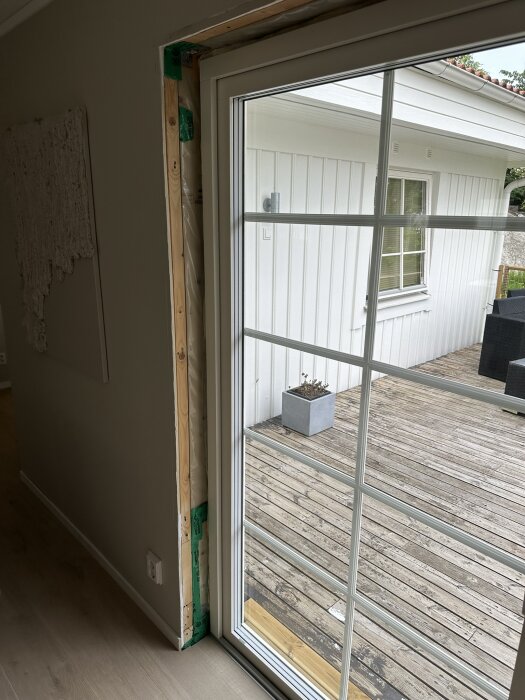 Nyligen installerad vit dörr med glasrutor sedd från insidan av vardagsrummet, öppning mot en träaltan och ett vitt hus i bakgrunden.