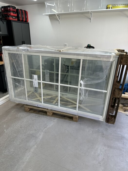 Fönster inplastade och placerade på en lastpall inomhus, redo för installation i ett byggprojekt. Verktyg och förvaring syns i bakgrunden.