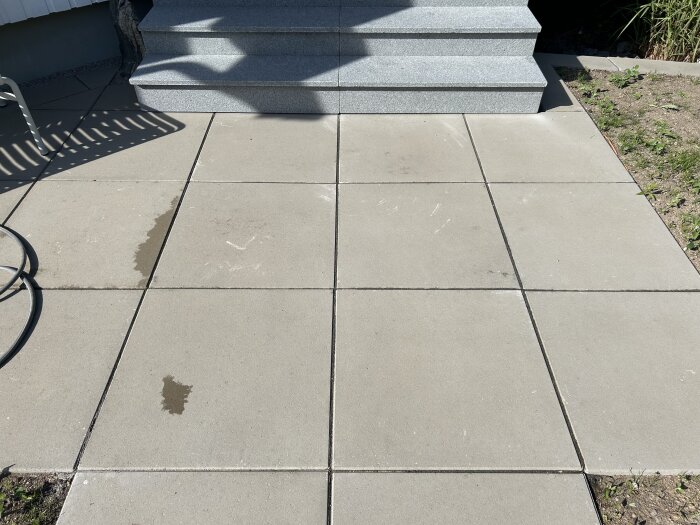 Betongplattor i ljusgrå färg, nyligen placerade med skrap- och fettfläckar synliga. Grå trappa av granit i bakgrunden och en del gräs syns på kanten.