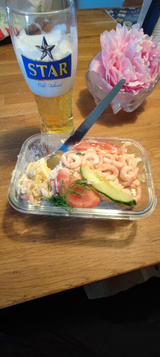 Smörgåstårta med räkor, avokado, tomat och dill på ett bord, en öl i ett glas märkt "STAR" och en vas med rosa blommor i bakgrunden.