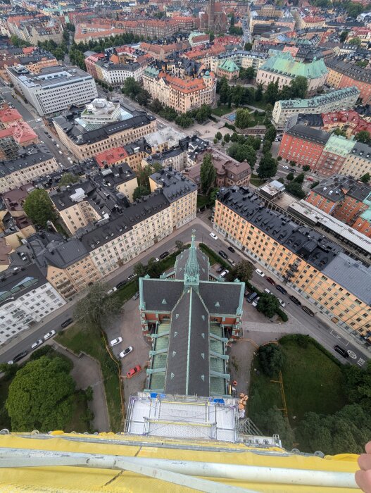 Utsikt från hög byggställning över stadsmiljö med färgglada byggnader, träd och kvadratiska innergårdar, samt taket på en historisk byggnad i förgrunden.