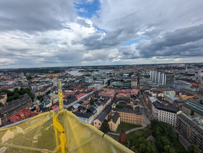 Utsikt över stad från en hög byggställning, med tak, kyrkor och olika byggnader under en delvis molnig himmel.