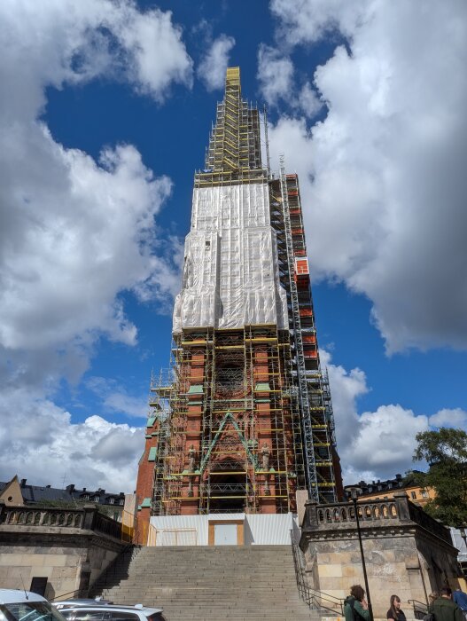 Byggställning runt en stor tegelbyggnad med stegar och plattformar, visar omfattande renovering under en klar blå himmel med moln.