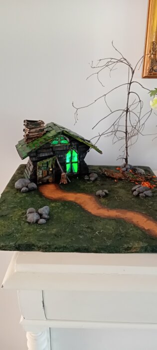 En miniatyrmodell av ett litet hus med grönt tak och lysande gröna fönster, omgiven av en stig, stenar, och ett träd på en grön yta.