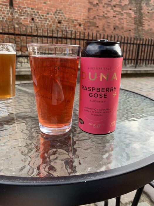 Ett glas och en burk hallonöl av varumärket Duna Raspberry Gose på ett glasbord, med en tegelvägg i bakgrunden.