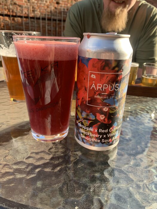 Ett glas röd öl och en ölburk från Ārpus Brewing Co. Pineapple x Red Currant x Blackberry x Vanilla Smoothie Sour Ale på ett utomhusbord med soligt väder.