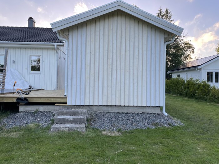 Två vita hus med svart tak. Ett litet platt tak på vänster sida och ett spetsigt tak på höger sida. Tät vegetation i bakgrunden.