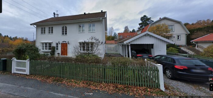 Ett hus med vit fasad och brunt tak, samt en vit fristående carport och två parkerade bilar. Grönmålade trästaket och höstlöv på marken.