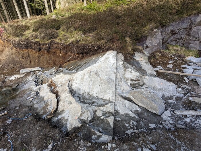 Bergvägg med borrade hål och sprickor från användning av snigeldynamit. Omgiven av jord och vegetationsklädd terräng i bakgrunden.