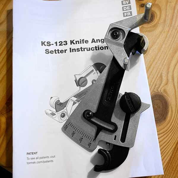 KS-123 knivvinkelinställare på ett instruktionshäfte, liggandes på ett träbord. Verktyget används för att enkelt ställa in och upprepa exakta vinklar.