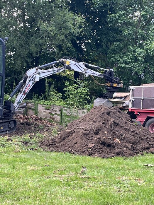 Grävmaskin vid rivning av pallkragar snett framför pallkragar, grushög i förgrunden på en gräsmatta. Lastbil syns i bakgrunden.