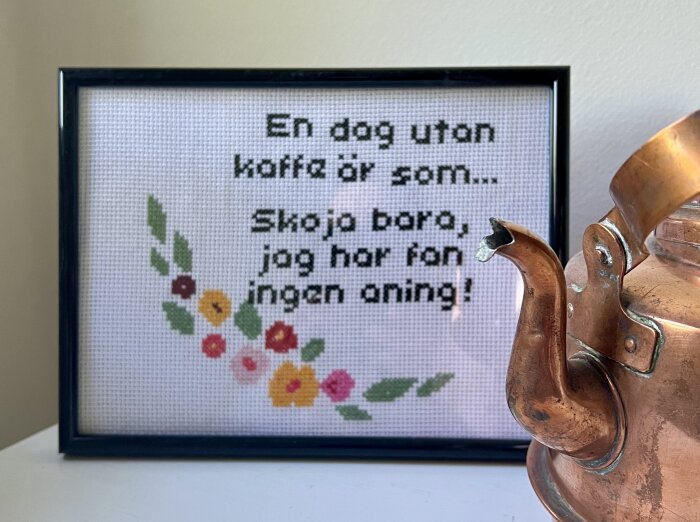 Korsstygnsbroderi i ram med texten "En dag utan kaffe är som... Skoja bara, jag har fan ingen aning!" bredvid en kopparkanna.