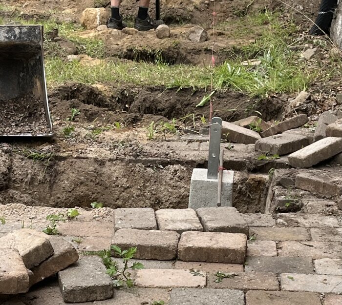 Utgrävda hål och platsen för en blivande balkong. En betongplint är delvis installerad i förgrunden, omgiven av stenar och gräs. Person i bakgrunden.