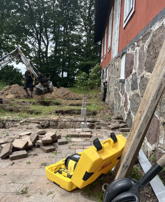 Grävmaskin och arbetare gräver plinthål vid röd murad byggnad med stenfundament, tegelstenar och verktygslåda utspridda i förgrunden.